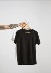 Tattooed biker hand holds wooden hangs with blank black and white t-shirts from premium thin cotton, isolated on white mockup