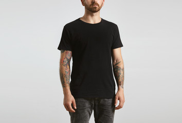 Brutal attractive bearded biker man with tattooed hands poses in black blank t-shirt from premium thin cotton, isolated on white mockup