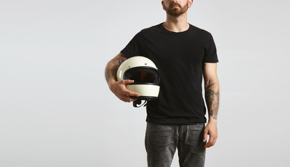 Brutal attractive bearded guy with tattooed hands poses in black blank t-shirt from premium thin cotton and holds ivory classic biker helmet, isolated on white mockup