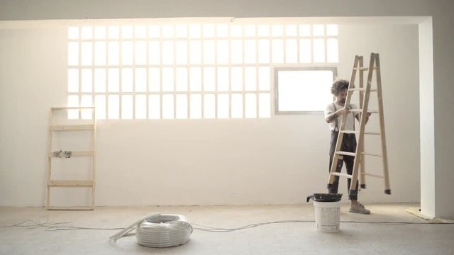 Funny curly man wears pants with suspenders brings wooden stairs, paints celling with white paint and takes a look at his work after Home diy renovation