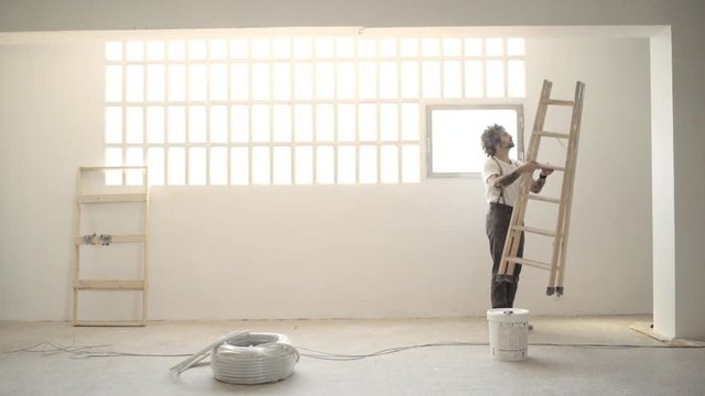 Funny curly man wears pants with suspenders brings wooden stairs and starts to paint celling with white paint Home diy renovation