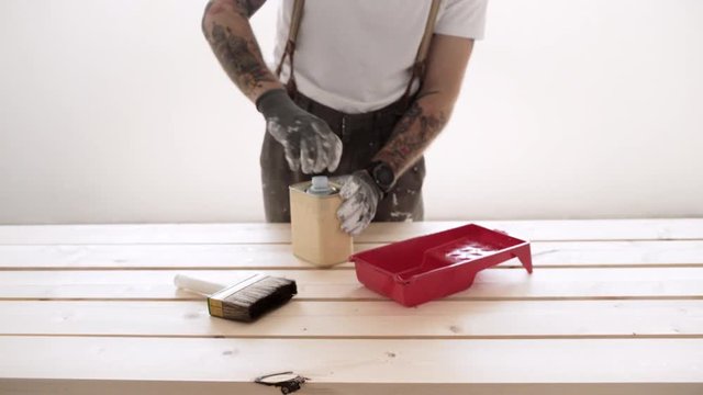 Unrecognizable man opens can with transparent wood oil and pours it in red container, prepares for work with wooden boards