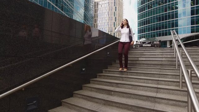 Beautiful girl is going down the modern stairs and talking on the phone near luxury offices and skyscrapers (steadicam shot)