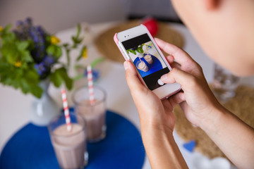 Woman taking photo of many glasses of fresh cold vanilla and chocolate smoothies with straws on table with flowers. Telephone photo of table setting for breakfast. Healthy snacks. Drinks for health.