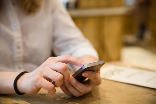 Close up photo of beautiful young woman sitting cozy at wooden table. Girl using  touch screen smartphone. Typing on the telephone.