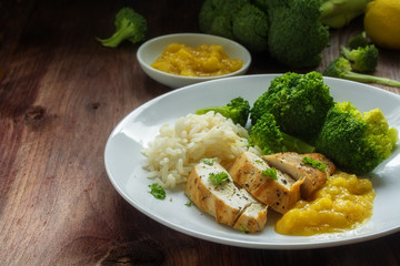 fried chicken breast fillet with stewed fruit sauce, broccoli an rice on a dark wooden table