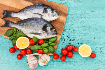 Fresh dorado fish on wooden cutting board, tomatoes, basil garlic and lemon on blue table. Top view, copy space 