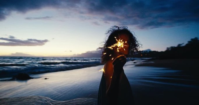 Beautiful young woman walking with sparkler fireworks on the beach at twilight