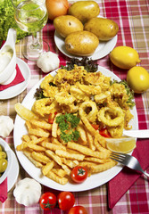Serving of breaded and deep-fried calamari and fries on a white plate served on a table with fresh vegetables, lemon and a glass of white wine.