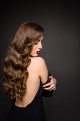 Naklejka premium Portrait of beautiful model lady with long brown hair posing over black background. Pretty woman with red lips showing her gorgeous back in studio. Skincare concept.