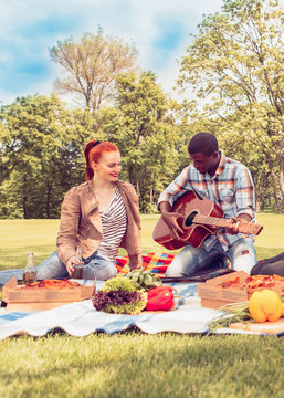 Picture of happy couple resting on picnic. Caucasian woman and Afro-American man sitting on picnic rug and playing guitar.