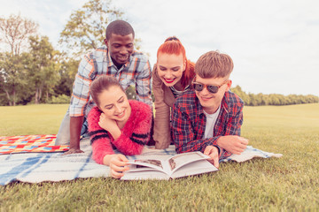 Four young friends picnicking in park. People lying on picnic rug and looking into book. Happy and cheerful people spending free time.