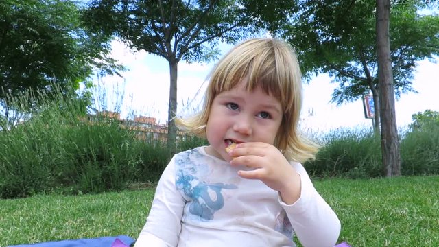 little blonde cute child sitting in grass at urban park eating cupcake, talking and laughing
