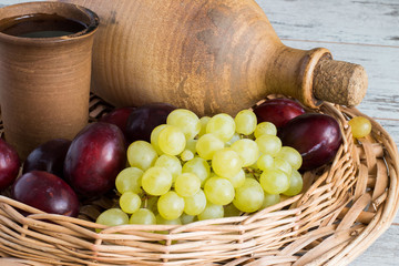     Clay bottle, a glass with wine, grapes and ripe plums.    Clay bottle, a glass with wine, grapes and ripe plums in a wicker basket on a light wooden table.