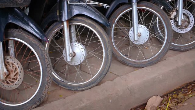 Forks, fenders and rusty spoked front wheels of many motorcycles, parked on a sidewalk along a typical street in Asia.