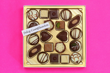 Feliz cumpleanos (Happy birthday in Spanish) card with box of assorted chocolates on pink background
