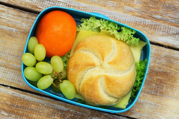 Lunch box containing cheese roll, mandarine and grapes on rustic wooden surface
