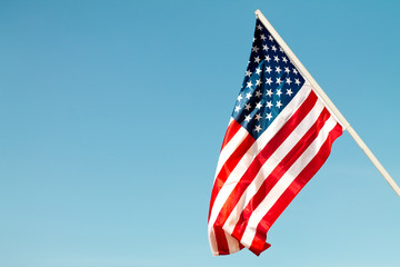 United States flag blows in the wind against a blue sky attached to the wall from the side. - obrazy, fototapety, plakaty