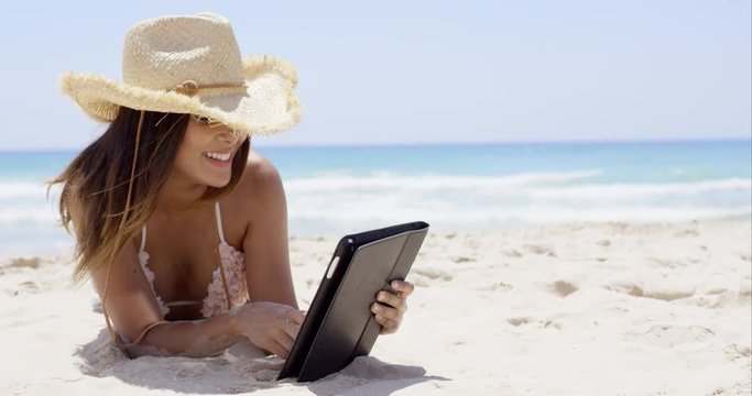 Beautiful woman on beach wearing straw hat works on her computer tablet as the sun pours down from above