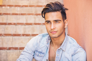 Portrait of handsome man with tattoo on arms sitting on cement stairs while leaning on wall, looking at camera
