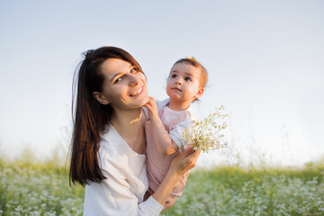 Beautiful portrait of mother embraces a daughter, toddler, baby, smiling with flowers in hand, on flower field, nature, sky background. Motherhood and childhood.