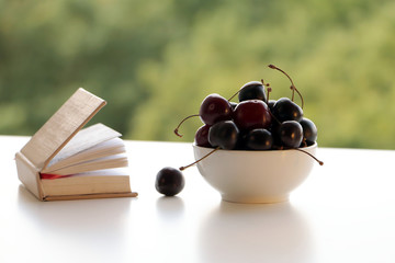 Cherry in a bowl on a white table with open book against summer green background. Selective focus.
