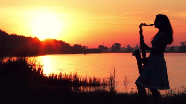 video silhouette of a young woman in a dress inspired by and quietly playing the saxophone at dawn on the river