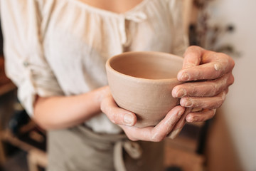 Potter woman keeping clay bowl in dirty palms closeup. Unrecognizable female craftsman with her creation at studio