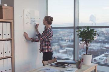 Businesswoman writing day plan on white magnet board, modern office. Side view of caucasian female...