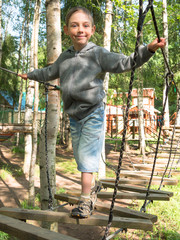 Little boy standing on a wooden ladder and holding the rope with both hands on the background of green forest