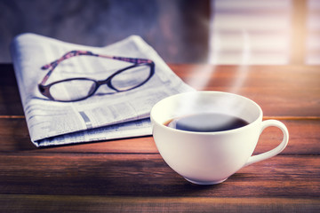 Coffee cup with newspaper and glasses in background on wooden table. Vintage tone photo with sunlight filter effect.