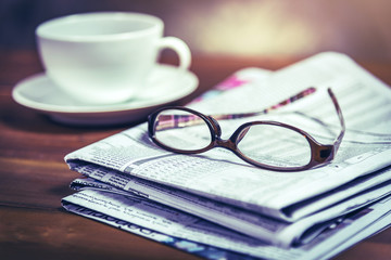 Fototapeta premium Glasses on pile of newspaper with coffee cup in background.Vintage tone photo with selective focus on glasses.