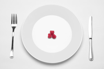 Healthy eating in the restaurant and diet Topic: white plate with three ripe raspberries and a metal knife and fork lying on a white table in the studio isolated top view