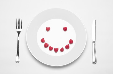 Healthy eating in the restaurant and diet Topic: white plate with raspberries and a metal knife and fork lying on a white table in the studio isolated top view berry smile