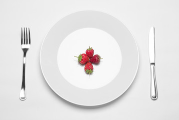 Healthy eating in the restaurant and diet Topic: white plate with three strawberries and a metal knife and fork lying on a white table in the studio isolated top view