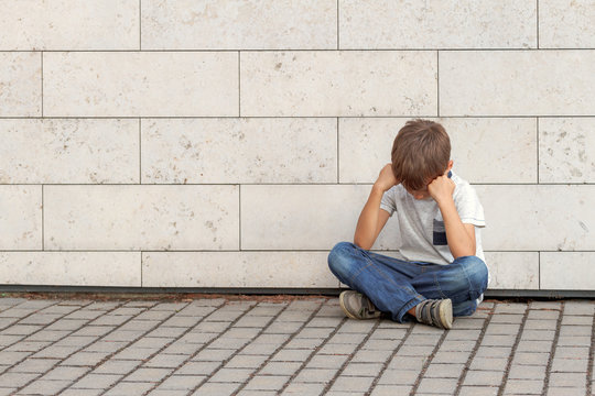 Sad, lonely, unhappy, upsed child sitting alone on the ground. Boy holding his head, look down. Outdoor