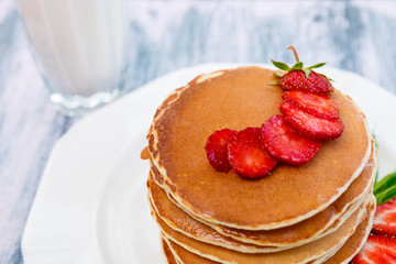 Closeup of pancakes with fresh strawberry and mint near glass with milk on white plate on pink wooden background. Stack of pancakes on the table.