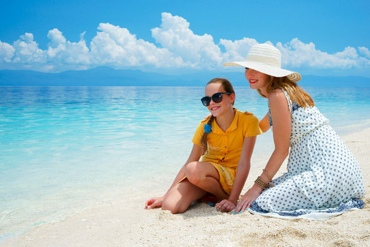 Youn europian woman in light dress is sitting on the white sand beach near beautiful tropical sea and cares about her teen daughter at sunny day