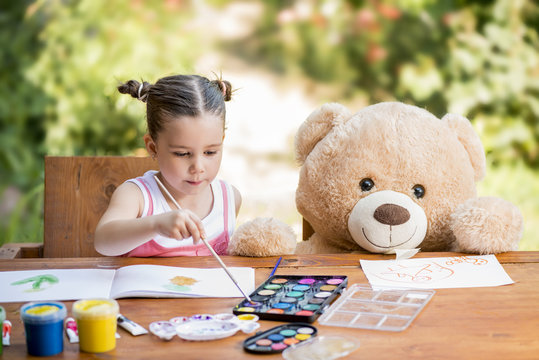 Adorable little girl painting outdoor in a sunny summer day with her teddy bear friend. Outdoor education concept