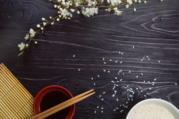 Sushi chopsticks over soy sauce bowl, rice and sakura blossom on a black background. 