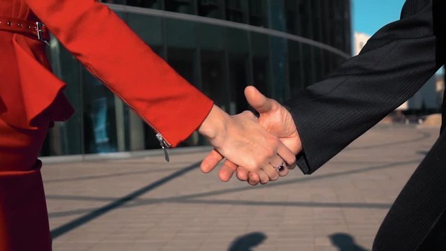 Two Business partners men in black suit and women in red bright dress do a hand-shake with glass business centre at the background. Slow motion close up.