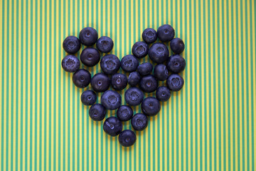 Many fresh blueberries arranged in the shape of heart on green-yellow stripe background