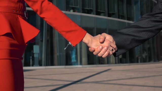 Two Business partners men in black suit and women in red bright dress doing a handshake with glass business centre at the background. Slow motion close up.