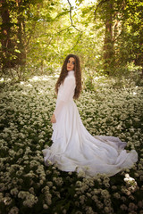 Obraz na płótnie Canvas Beautiful woman in long white dress standing in a forest on a carpet of flowers