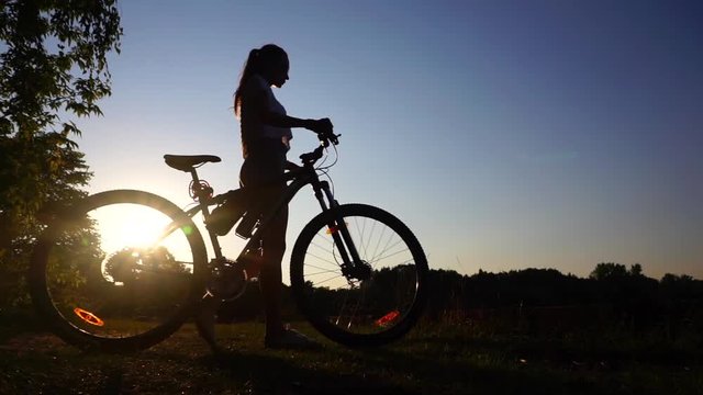 Silhouette of girl cyclist with ponytail hairstyle walking with her bike. Slow motion shot against sunset