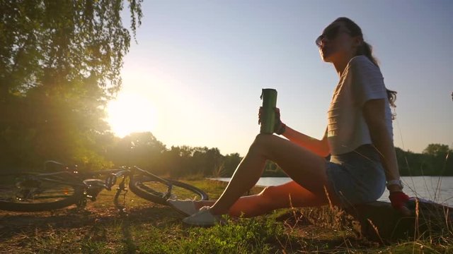 Slender girl cyclist with ponytail hairstyle drinking from her bottle. Slow motion shot against sun