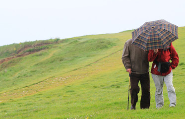 An elderly couple down the hill in the rain, holding an umbrella.