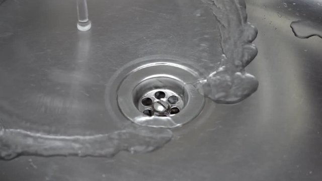 The stream of water falls from a tap in the sink in stainless steel and goes down the drain. Slow motion, high speed camera, 250fps
