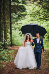 Beautiful bride and groom walking under umbrella at park. Bride with red hair. Pine forest. Newlyweds walking.