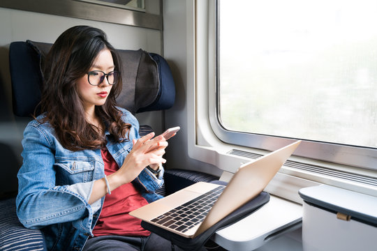 Cute Asian woman using smartphone and laptop on train, copy space on window, business travel or technology concept